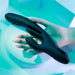 A hand holds the Playboy Rapid Rabbit Thrusting Vibrator with Flapping Shaft, a sleek black personal massager ideal for g-spot stimulation. It is set against a soft blue background with geometric shapes, enhanced by gentle lighting for a serene ambiance.