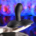 The Playboy Sittin' & Spinnin' Hands Free G-Spot Vibrating Grinder Pad, a black, curved pleasure toy with a textured base, is shown on a white mannequin hand against a vibrant background of purple, blue, and red hues.