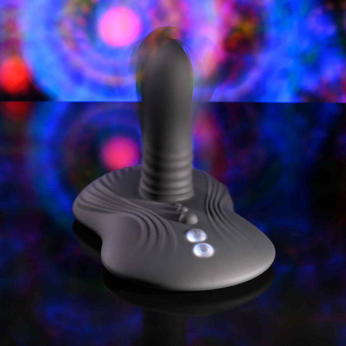 The Playboy Sittin' & Spinnin' Hands Free G-Spot Vibrating Grinder Pad, a sleek gray silicone toy with buttons and a curved, ribbed design, stands upright on a reflective surface amid swirling colorful lights and an abstract background.