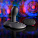 The Playboy Sittin' & Spinnin' Hands Free G-Spot Vibrating Grinder Pad, a black silicone toy with a rounded, textured shaft and remote control, rests on a reflective surface with colorful, blurred lights in the background.