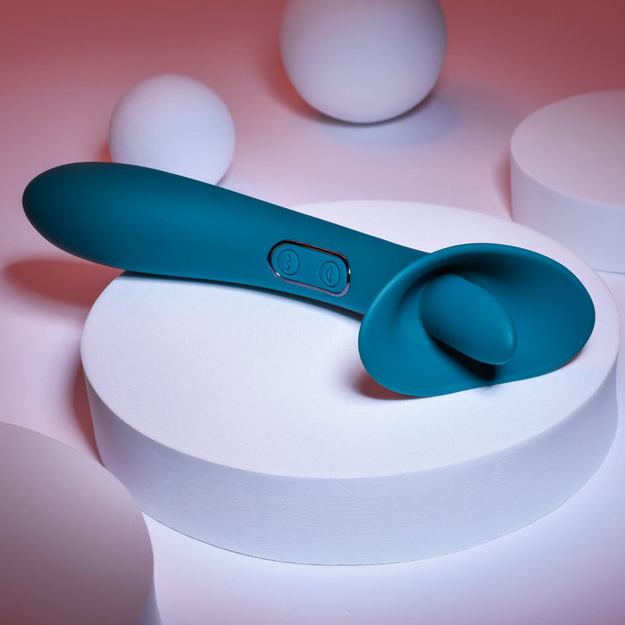 The Playboy True Indulgence Dual Ended Flicking Tongue Vibrator - Green by Evolved Novelties, boasting a smooth premium silicone finish and dual control buttons on its handle, is showcased on a white cylindrical pedestal. The backdrop features spherical shapes with a striking red gradient lighting effect.