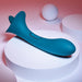 A green Playboy True Indulgence Dual Ended Flicking Tongue Vibrator by Evolved Novelties, ergonomically designed and made of premium silicone, is displayed on a white circular platform against a pink and white background. The toy features a curved shape and two buttons near its base. Round objects are placed in the background with soft, diffused lighting.