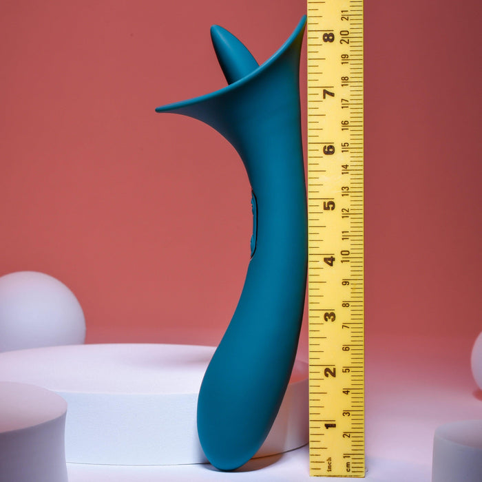 The Playboy True Indulgence Dual Ended Flicking Tongue Vibrator in green from Evolved Novelties, made from premium silicone with an ergonomic design and flared top, stands upright next to a yellow ruler on a pink background, measuring approximately 9 inches. The scene includes white cylindrical objects of varying heights in the background.