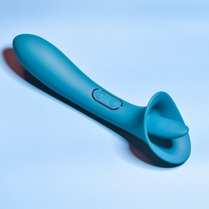 A green Playboy True Indulgence Dual Ended Flicking Tongue Vibrator from Evolved Novelties lies on a light blue surface. The device boasts an ergonomically designed, smooth cylindrical handle with two intuitive buttons and a uniquely shaped, flared end for targeted use. It’s waterproof, ensuring versatile relaxation.