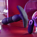 The Playboy Trust the Thrust Vibrating & Thrusting Anal Plug with Remote by Evolved Novelties, featuring a ribbed, dark-colored design and battery operation, is displayed on a red surface. The image is highlighted with purple and pink lighting, while circular transparent objects are seen in the background.