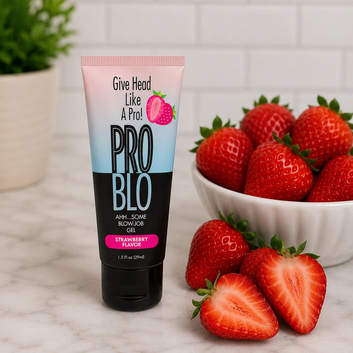 A tube of Pro Blo Oral Pleasure Gel 1.5 oz (strawberry flavor, one of 8 available) stands upright on marble beside a bowl of fresh strawberries and a cut strawberry, with a potted plant blurred in the background.