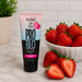 A tube of Pro Blo Oral Pleasure Gel 1.5 oz (strawberry flavor, one of 8 available) stands upright on marble beside a bowl of fresh strawberries and a cut strawberry, with a potted plant blurred in the background.