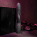 A sleek, black Rabbit Hole Double Ended G-Spot and Clitoral Suction Rabbit Vibrator from Evolved Novelties stands upright on a dark surface with smooth, round stones in the background. This silicone device features dual prongs and a rounded, textured head designed for both g-spot and clitoral stimulation. Buttons for settings are conveniently located near the base of the toy. The overall ambiance is dim and intimate.
