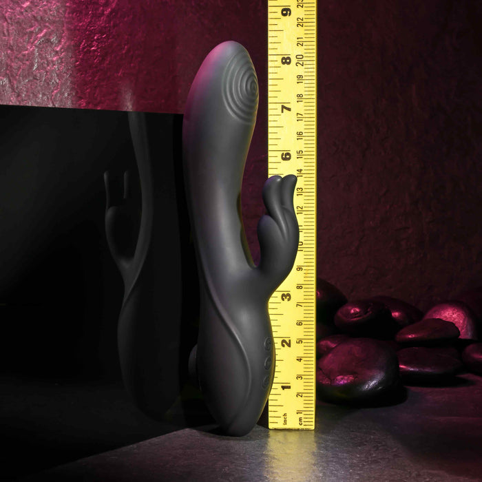 The Rabbit Hole Double Ended G-Spot and Clitoral Suction Rabbit Vibrator by Evolved Novelties, a black, curved adult toy made of premium silicone with dual prongs, stands upright against a yellow measuring tape showing a height of approximately 8 inches. The background features a dark, stone-like surface with round black stones and a pinkish light, hinting at g-spot and clitoral stimulation.