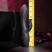 The Rabbit Hole Double Ended G-Spot and Clitoral Suction Rabbit Vibrator by Evolved Novelties, a black, curved adult toy made of premium silicone with dual prongs, stands upright against a yellow measuring tape showing a height of approximately 8 inches. The background features a dark, stone-like surface with round black stones and a pinkish light, hinting at g-spot and clitoral stimulation.