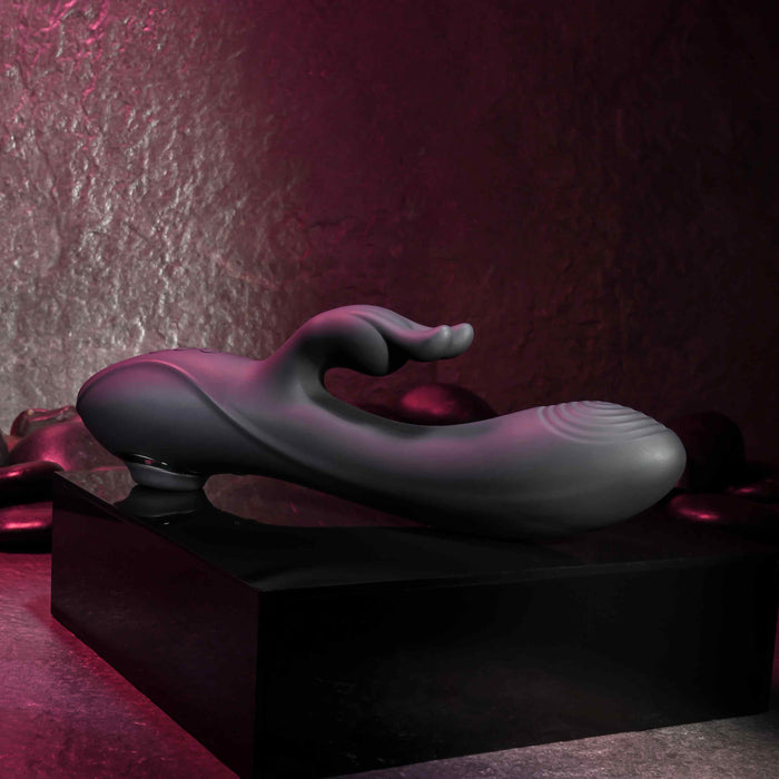 The Rabbit Hole Double Ended G-Spot and Clitoral Suction Rabbit Vibrator by Evolved Novelties is a modern, sleek black abstract sculpture with smooth curves and a flowing shape, displayed elegantly on a black rectangular pedestal. Crafted from premium silicone, its textured background is illuminated with subtle pink and purple lighting, creating an elegant and mysterious ambiance.