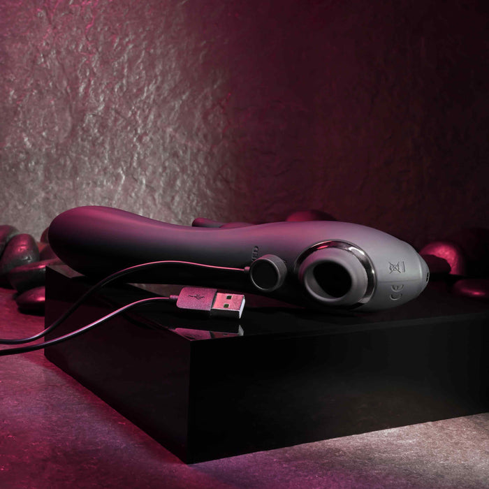A black, ergonomically designed Rabbit Hole Double Ended G-Spot and Clitoral Suction Rabbit Vibrator by Evolved Novelties rests on a shiny black platform. Made of premium silicone, it features a smooth texture with an attached USB charging cable, promising powerful motors for clitoral and g-spot stimulation. The background is dimly lit with rock-like textures bathed in red-tinted light.