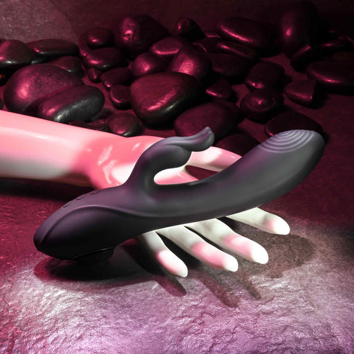 A black Rabbit Hole Double Ended G-Spot and Clitoral Suction Rabbit Vibrator from Evolved Novelties, crafted from premium silicone with a dual-prong design, rests on a pale mannequin hand. The background, featuring smooth dark stones and bathed in pink and purple lighting, creates a sensual atmosphere perfect for g-spot stimulation.