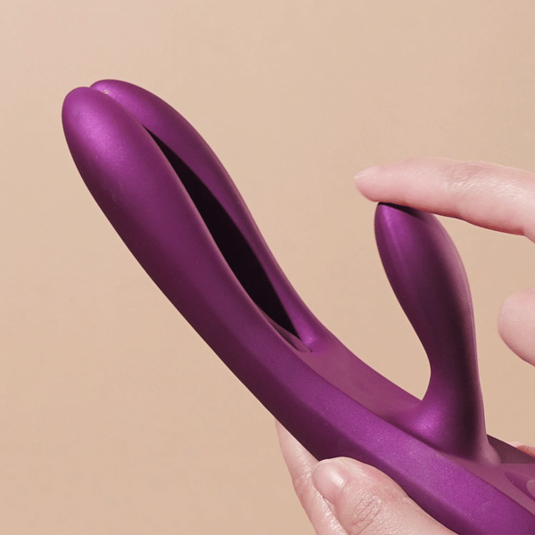 A hand holds the Terri App-Controlled G-Spot Tapping Rabbit Vibrator in purple, featuring a curved, split design and a small extension for G-spot stimulation, set against a beige background.