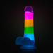 The Blush Rainbow Rave Glow in the Dark 6.5 Inch Silicone Dildo stands upright against a dark background, highlighting its playful and vibrant multicolored design.