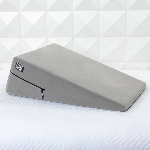 A gray Liberator Ramp Sex Positioning Cushion - 30 Inches with a zippered cover rests on a white quilted surface in front of a geometric-patterned white wall.