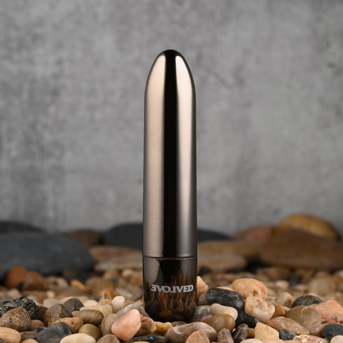 The Real Simple Rechargeable Waterproof Bullet Vibrator in shiny black chrome stands upright on smooth pebbles, with "EVOLVED" appearing near its base and a concrete wall in the background.