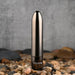 The Real Simple Rechargeable Waterproof Bullet Vibrator in shiny black chrome stands upright on smooth pebbles, with "EVOLVED" appearing near its base and a concrete wall in the background.