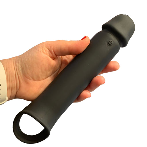 A hand with red nail polish holds an NS Novelties Renegade Brute Vibrating Hollow Black Penis Extension with Remote, which is cylindrical with a rounded tip and features a loop handle at its base. A visible power button on the side near the rounded end suggests it has a vibrating shaft. The person is wearing a light-colored wristband.
