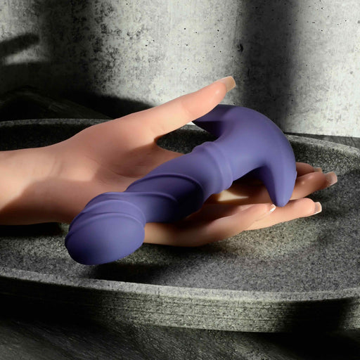 A hand with manicured nails holds the purple Ring It Remote Control Rimming Vibrating Prostate Plug, featuring a curved handle, resting in a gray stone dish against a concrete background.