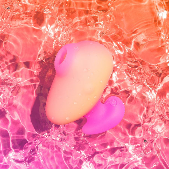 The Romp Kiss Beginner Air Pulsation Clitoral Stimulator, in a gradient of pink and orange silicone, features an ergonomic design on a rippling pink water surface. Water droplets enhance its vibrant, playful aesthetic, ideal for travel size convenience.