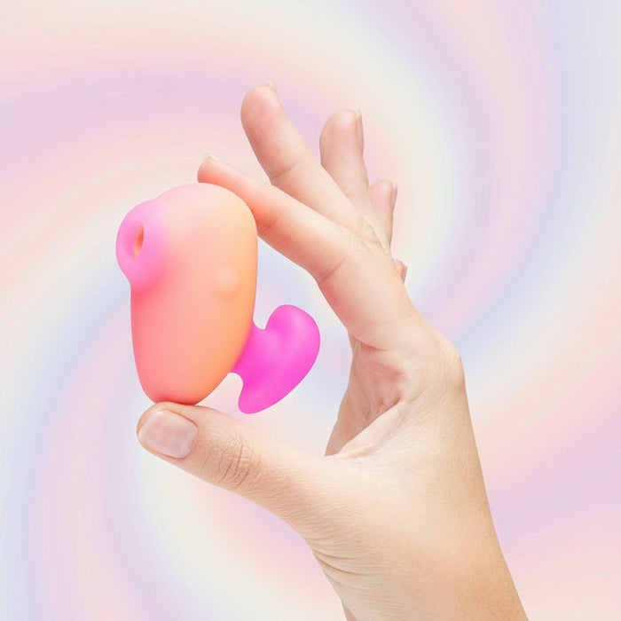 A hand holds the Romp Kiss Beginner Air Pulsation Clitoral Stimulator, a small, pink and orange, soft-textured device with a curved shape against a dark teal background, perfect for travel convenience.