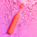 The Romp Pop Pinpoint Clitoral Stimulator with 2 Heads, in coral, features water droplets on its surface as it rests on rippling water reflecting pink light.