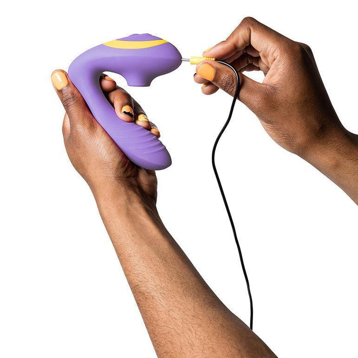 Someone holds the Romp Reverb Pleasure Air Vibrator for G-Spot & Clitoris in one hand and plugs in its charging cable with the other, set against a white background.