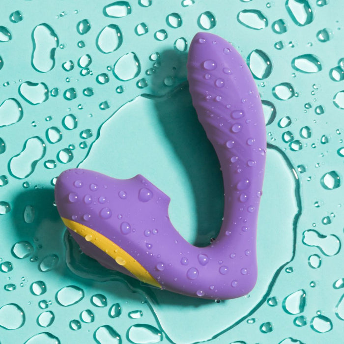 The Romp Reverb Pleasure Air Vibrator for G-Spot & Clitoris, in purple and yellow with a curved waterproof design, rests on a teal surface with water droplets, ready to provide intense stimulation.