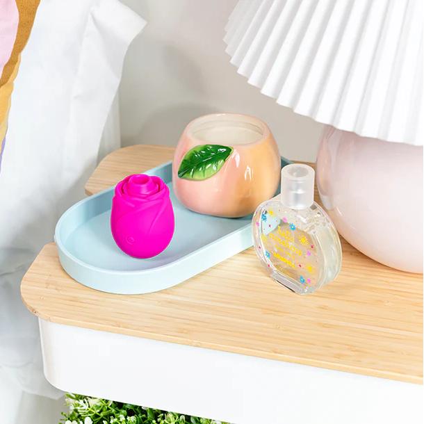 On a bedside table sits the Romp Rose 2 Pleasure Air Clitoris Stimulator, a pink silicone device, along with a peach candle adorned with a green leaf and a clear perfume bottle. Nearby is a white lamp with a pleated shade.