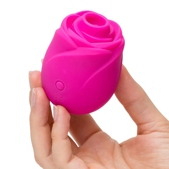 A hand holds the Romp Rose 2 Pleasure Air Clitoris Stimulator, a small pink silicone device with a button on the side, designed for gentle clitoral stimulation.