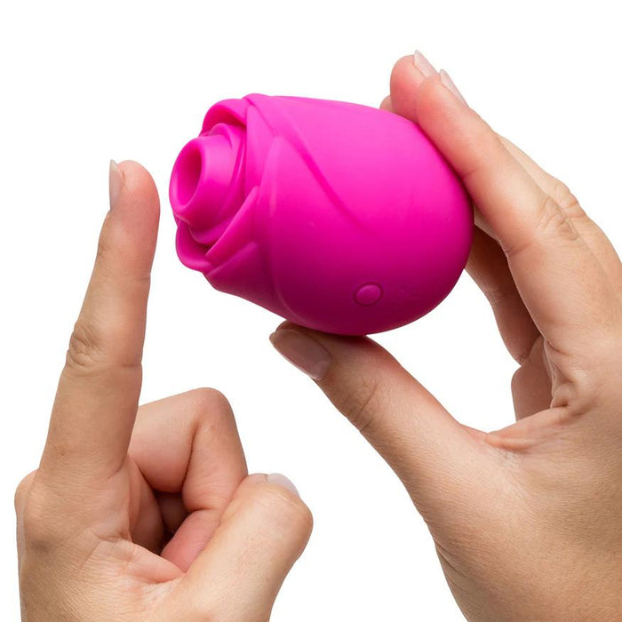 A person holds the Romp Rose 2 Pleasure Air Clitoris Stimulator—a pink, rose-shaped silicone toy—between their hands, pointing toward its opening. The white background highlights its design for clitoral stimulation.