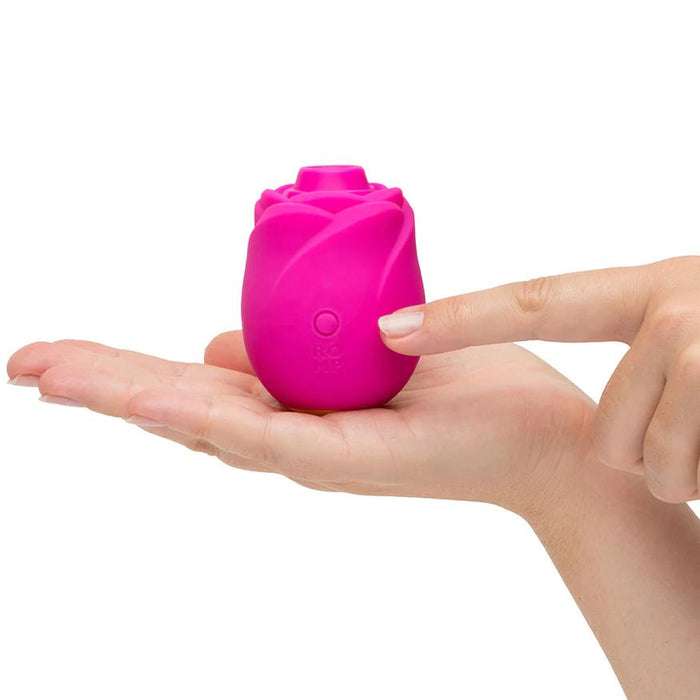 A person holds the Romp Rose 2 Pleasure Air Clitoris Stimulator, a compact pink rose-shaped silicone toy, in their palm and presses its button for clitoral stimulation.