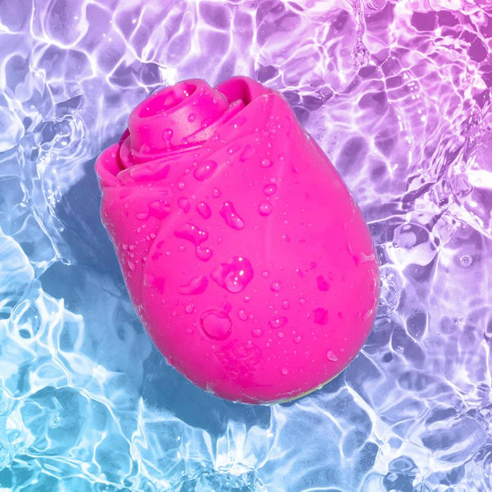 The Romp Rose 2 Pleasure Air Clitoris Stimulator—a bright pink, rose-shaped silicone toy—floats with water droplets on a blue and purple gradient background, hinting at innovative clitoral stimulation.
