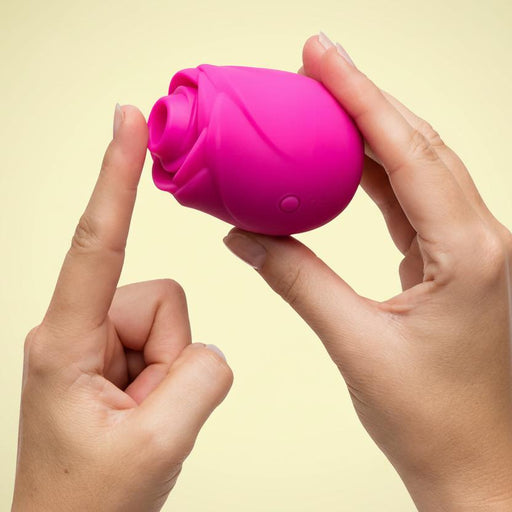 Two hands hold the Romp Rose 2 Pleasure Air Clitoris Stimulator, a small pink rose-shaped silicone device with a button, as one finger points to its opening; it stands out against a plain light yellow background.