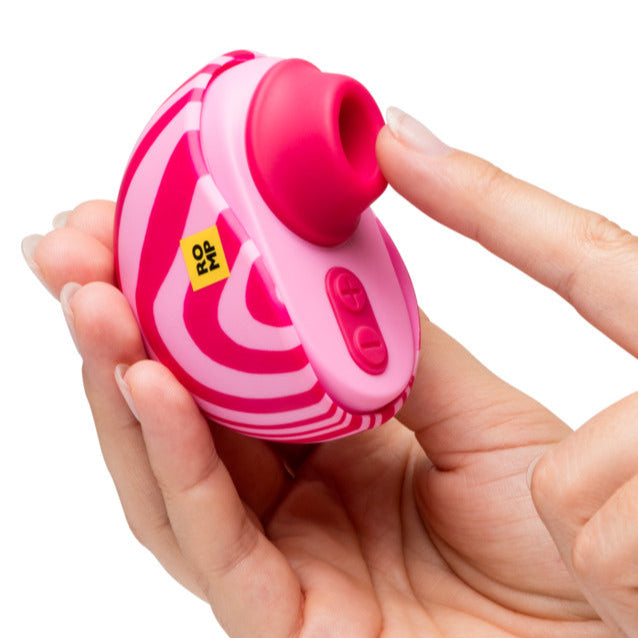 A hand holds the Romp Spin Small Discreet Pleasure Air Clitoral Stimulator, a pink and red travel-sized vibrator with a circular opening and control buttons, while another hand points toward the opening.