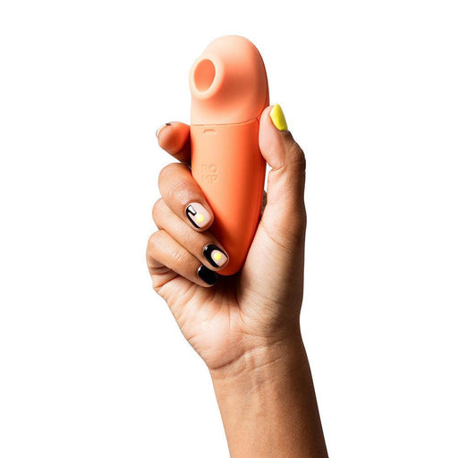 A hand with painted nails is holding an orange, compact, and ergonomically designed personal wellness device against a white background. The Romp Switch X Beginner's Pleasure Air Clitoral Stimulator by WOW features Pleasure Air Technology and offers 6 intensity levels. The device has a rounded top with an opening and displays the brand name "ROMP" on its body.
