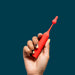 A hand with black and yellow nails holds the Romp Pop Pinpoint Clitoral Stimulator with 2 Heads—a bright red, waterproof silicone vibrator—against a solid teal background.