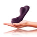 A hand is holding a dark purple, ergonomically designed Ruby Glow Ride On External Hands-Free Humping Vibrator from Rocks Off. The non-penetrative massager features a streamlined design with smooth texture and curves, set against a white background.