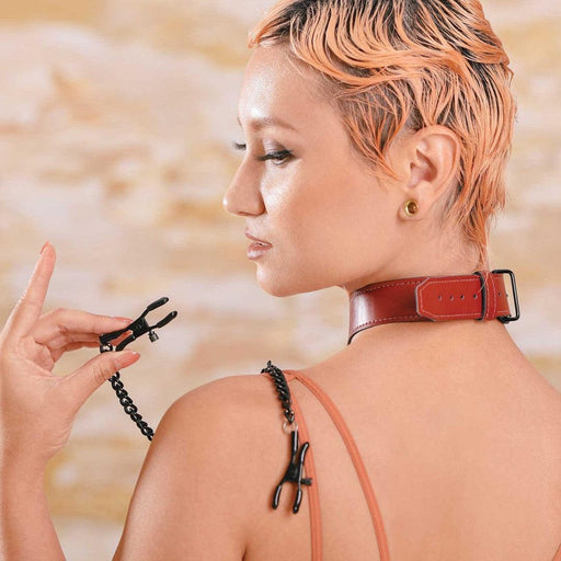 A woman with a trendy short haircut and a **Sportsheets** saffron collar with nipple clamps seen from the back as she glances over her shoulder, holdingthe saffron nipple clamp