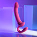 The Sharing Is Caring Double Ended Wearable Strapless Strap On Vibrator in pink by Evolved Novelties, complete with a curved design, bulbous head, and a C-shaped handle, rests against a geometric, translucent blue statue. This waterproof delight boasts ten vibrating speeds and is set against a backdrop of blue and purple hues, casting a soft ambient glow.