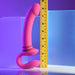 The Sharing Is Caring Double Ended Wearable Strapless Strap On Vibrator by Evolved Novelties, featuring a curved pink shaft and dual ends with 10 vibrating speeds, stands upright next to a yellow measuring tape. Set against a gradient of blue and purple hues, this versatile double-ended vibe promises excitement.