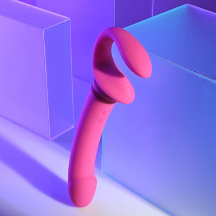 A pink, curved silicone sex toy from Evolved Novelties stands upright on a light surface. The background features translucent geometric shapes illuminated by blue and purple lights, creating a soft, futuristic aesthetic. The Sharing Is Caring Double Ended Wearable Strapless Strap On Vibrator is waterproof and boasts 10 vibrating speeds for diverse pleasure experiences.