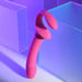A pink, curved silicone sex toy from Evolved Novelties stands upright on a light surface. The background features translucent geometric shapes illuminated by blue and purple lights, creating a soft, futuristic aesthetic. The Sharing Is Caring Double Ended Wearable Strapless Strap On Vibrator is waterproof and boasts 10 vibrating speeds for diverse pleasure experiences.