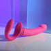 The Sharing Is Caring Double Ended Wearable Strapless Strap On Vibrator by Evolved Novelties is showcased on a reflective surface against a modern, abstract setting of transparent, blue-tinted panels. This pink, flexible silicone sex toy features ribbed detailing near the base and offers 10 vibrating speeds for endless pleasure.