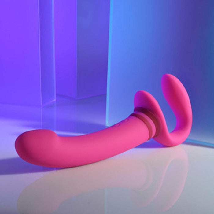 The Sharing Is Caring Double Ended Wearable Strapless Strap On Vibrator by Evolved Novelties, in a pink hue, is displayed on a smooth surface. Its curved shaft and attached smaller, curved end are showcased against a backdrop of blue and purple lighting with reflective surfaces, emphasizing its 10 vibrating speeds and waterproof design.