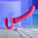 The Evolved Novelties' Sharing Is Caring Double Ended Wearable Strapless Strap On Vibrator in pink, a rechargeable adult toy with dual stimulators made from phthalate-free silicone and featuring an attached USB charging cable, is placed against a background with blue and purple hues. The waterproof toy, showcasing a sleek and modern design, is set on a reflective surface.
