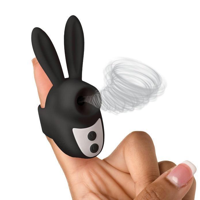 A person's hand is holding the Shegasm Sucky Bunny Beginner Clit Stimulator from XR Brands, a small black device shaped like a bunny's head with two long ears. Made of body-safe silicone, it features two buttons on the front and emits a swirling, wind-like effect from a circular opening on its face. The background is white.
