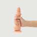 A hand holding the Sliding Skin Realistic Small 7 Inch Vanilla Silicone Dildo with Suction Cup from Lovely Planet, featuring a flesh-colored, lifelike design and a flared base. The background is plain and light gray.