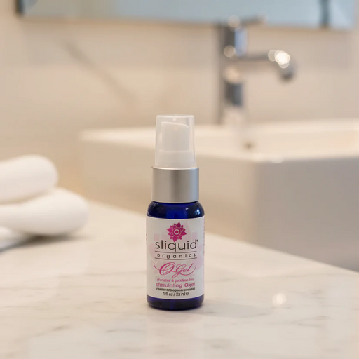 A 1 oz bottle of Sliquid Organics Stimulating Clitoral Orgasm Gel rests on a bathroom counter, with a sink, faucet, and neatly folded white towels blurred in the background.